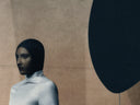 Person wearing a black hood against a beige wall with a large black circle.